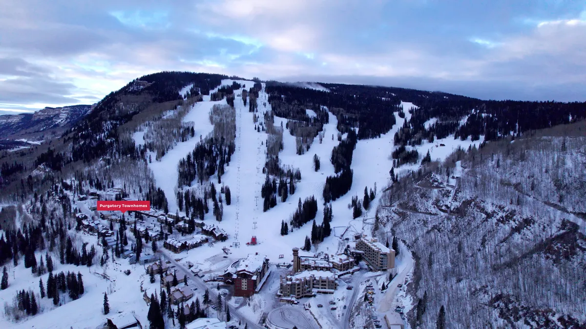 A wintry exterior view featuring ski slopes in the background, snow-covered trees, and a row of townhouses nestled at the base of the mountain. The atmosphere is lively, inviting, and perfectly captures the immediate proximity to the slopes and the resort's mountain charm.