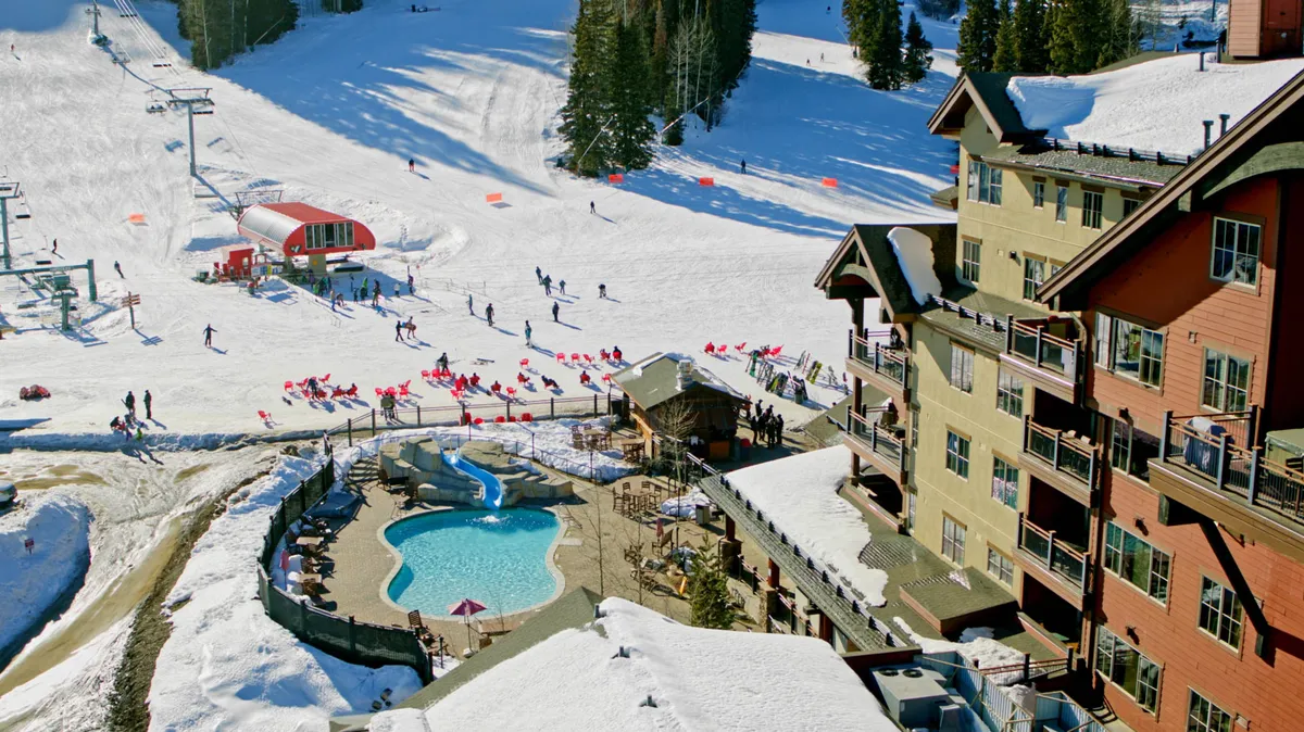 This exterior image captures the lively pool area at the base of the ski slopes, with skiers and snowboarders enjoying the snow and people gathered around the pool. The lodge buildings frame the scene, creating a festive, communal atmosphere that highlights the blend of mountain adventure and leisure.