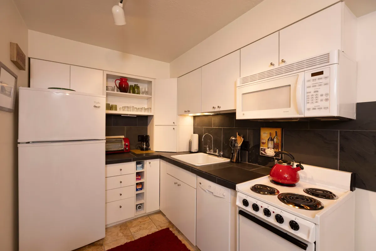 A close-up view of the kitchen’s red kettle on black countertops, surrounded by white cabinets and a stainless steel sink, highlighting the space’s inviting atmosphere for cooking and sharing meals.
