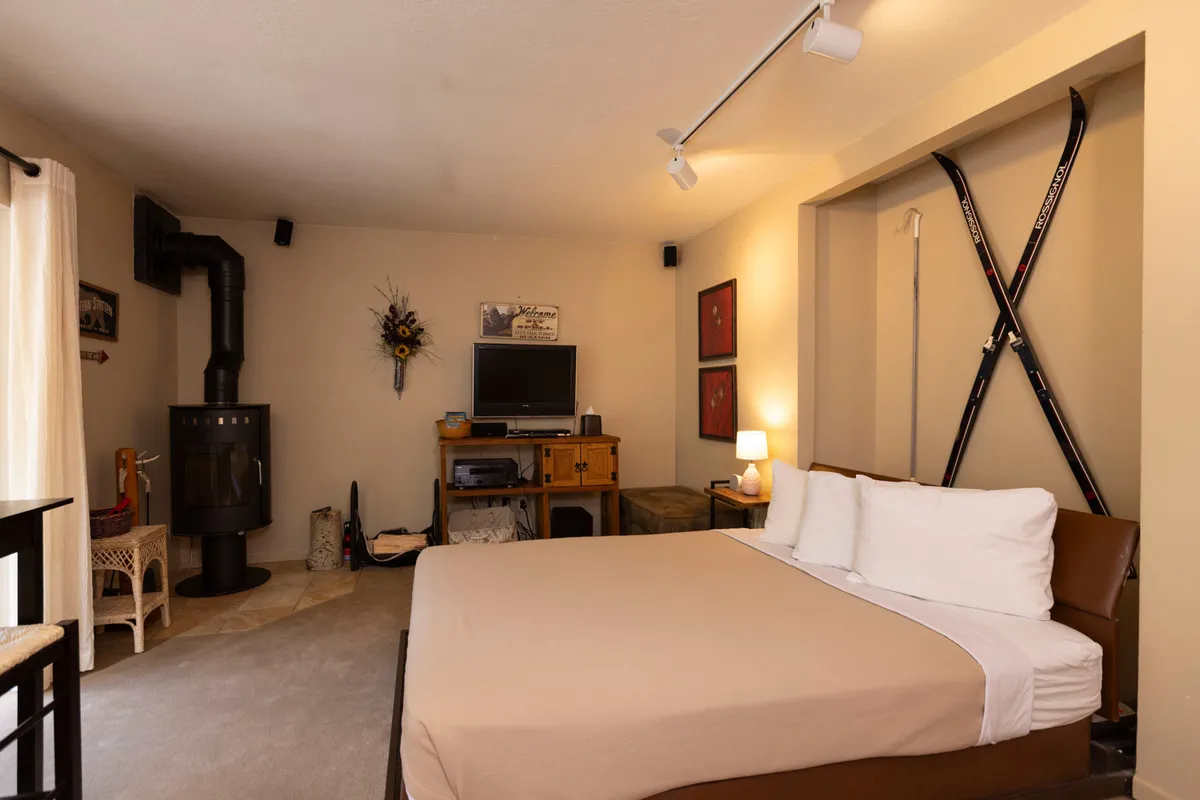 This cozy studio interior features a comfortable bed with crisp white linens, a wooden dresser, a sleek black stove for warmth, decorative mountain-themed wall art, and a pair of vintage skis mounted on the wall. The inviting atmosphere suggests relaxing evenings, whether enjoying a movie night, reading by the fire, or sipping hot cocoa after a day on the slopes.