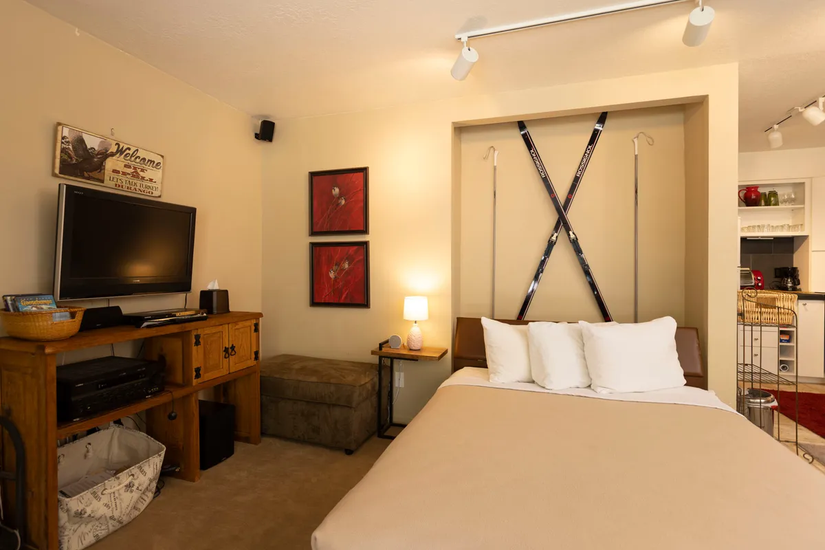 A welcoming studio interior with a bed dressed in crisp white pillows, a wooden dresser, and framed artwork on the wall. Ski equipment adds a touch of local character. The mood is relaxed, and the setup is ideal for unwinding after a day of mountain adventures or transitioning the space into a restful sleeping area.
