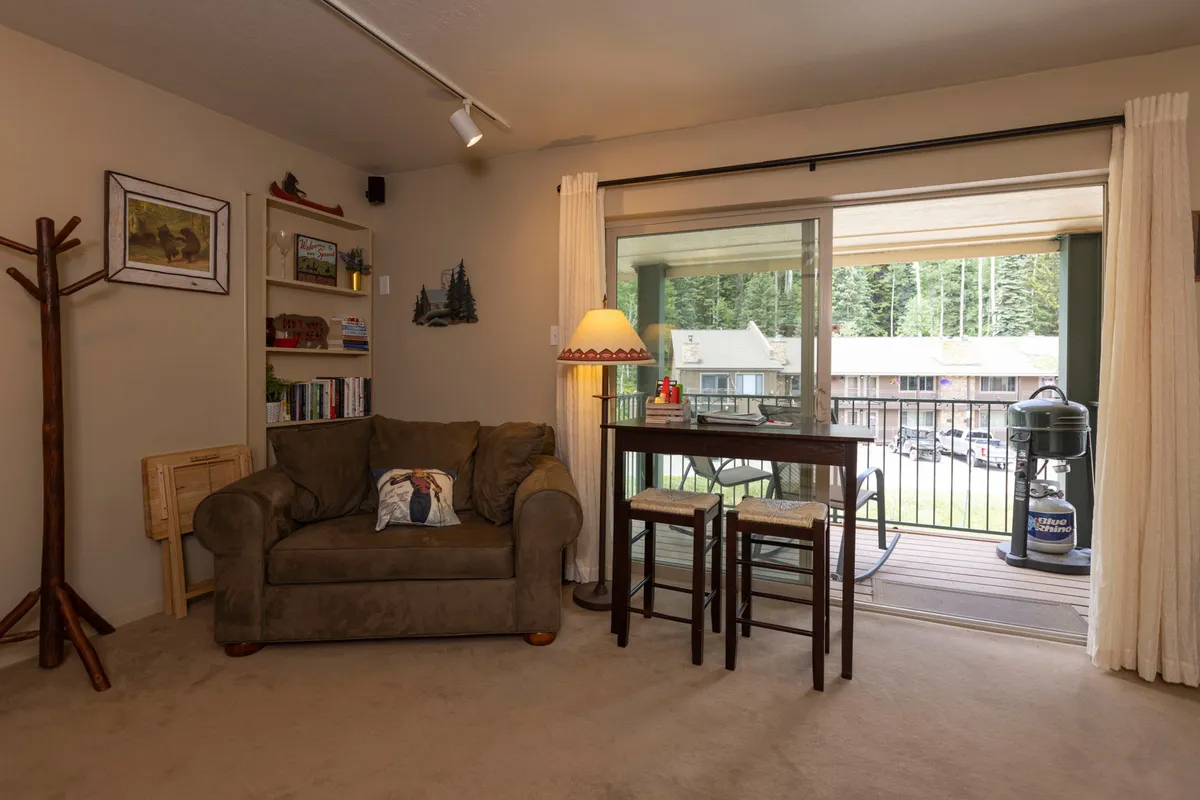 A comfortable studio living space featuring a cozy sofa with a decorative pillow, a small dining table with stools, and a large sliding glass door that leads out to the balcony. There is a warm lamp with a shade, and the room is bathed in natural light, enhancing the inviting atmosphere. The space highlights flexibility and relaxation, perfectly capturing the studio’s dual purpose as a living and lounging area.