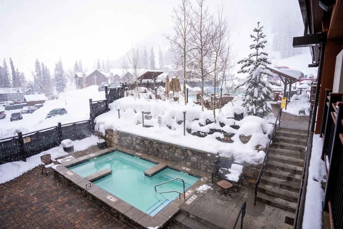 An inviting outdoor hot tub is nestled among a snowy landscape, with stepping stones leading down to the steaming water. The tranquil, secluded setting invites guests to soak and unwind while surrounded by stunning winter scenery, suggesting both relaxation and social connection.