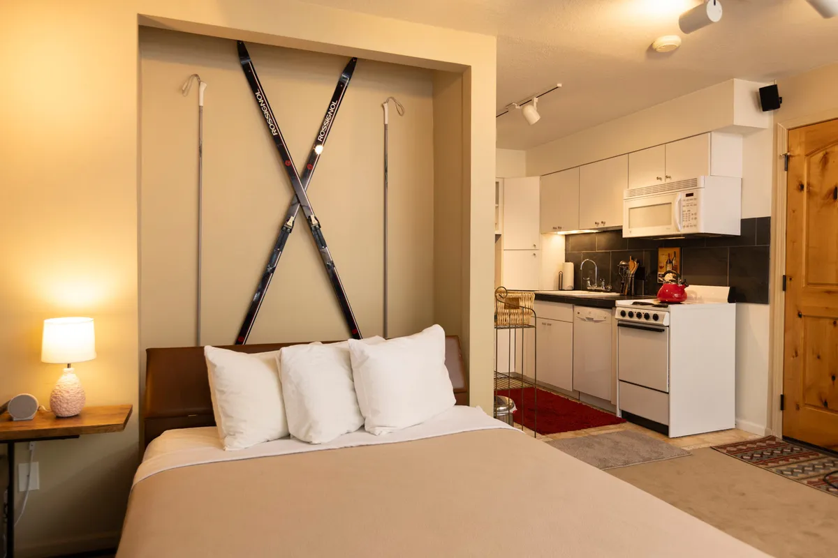 An inviting studio interior featuring a bed with plush white pillows, ski equipment mounted on the wall as decor, and a compact kitchen area. The layout demonstrates space-saving efficiency and warmth, ideal for small groups or couples. Hints of a mountain view appear through the window, reinforcing the connection to the alpine environment.