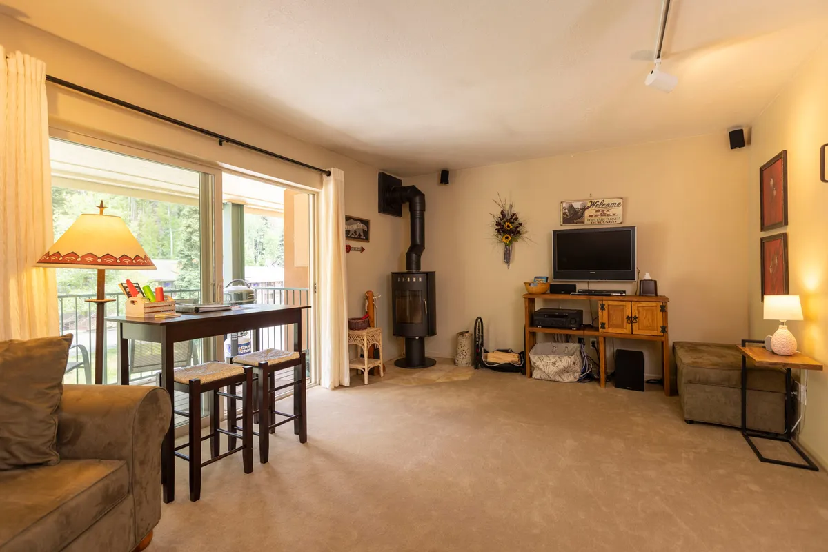 A warm, inviting studio corner with a cozy sofa, wooden table with bar stools, and a television set atop a modern stove. Decorative wall art completes the homely scene, creating a perfect spot for socializing, reading, or enjoying meals. The layout demonstrates how the compact space accommodates multiple functions seamlessly.