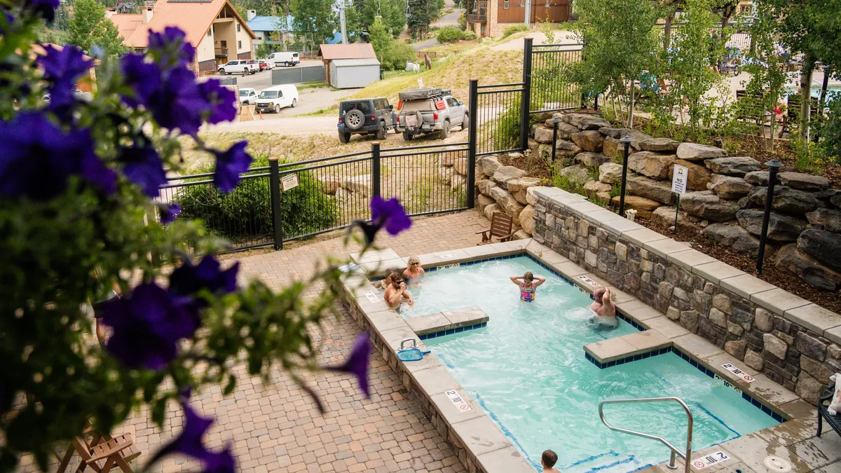 A sunlit swimming pool is ringed by lush greenery and a fence, with children playing and adults relaxing poolside. The joyful, communal mood highlights opportunities for socializing and family fun in the pool area.